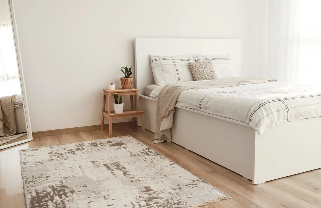 A neutral patterned area rug on a wood floor beside a low white bed and step-stool nightstand