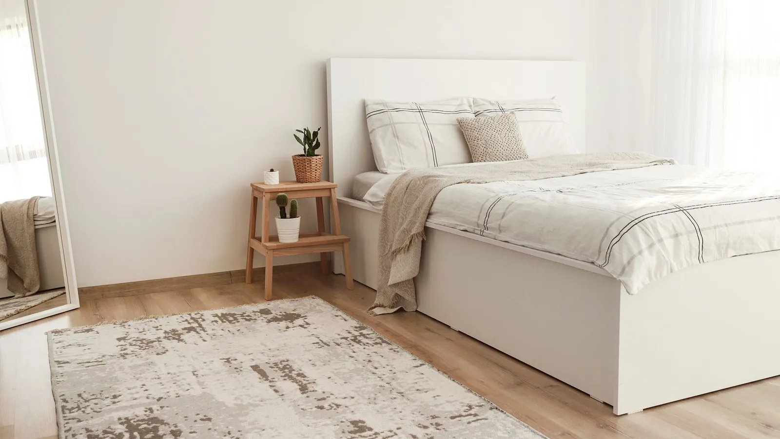 A neutral patterned area rug on a wood floor beside a low white bed and step-stool nightstand