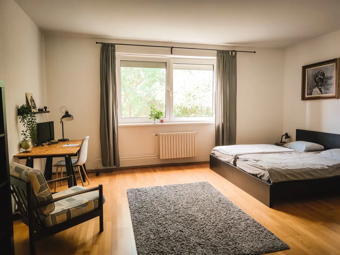 A bedroom with a large grey area rug, wooden desk, and armchair beside a window