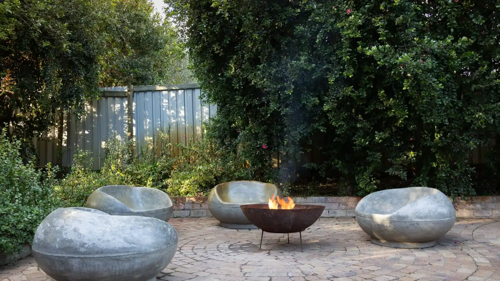 A lit corten steel fire bowl on a reclaimed-brick paver patio surrounded by greenery
