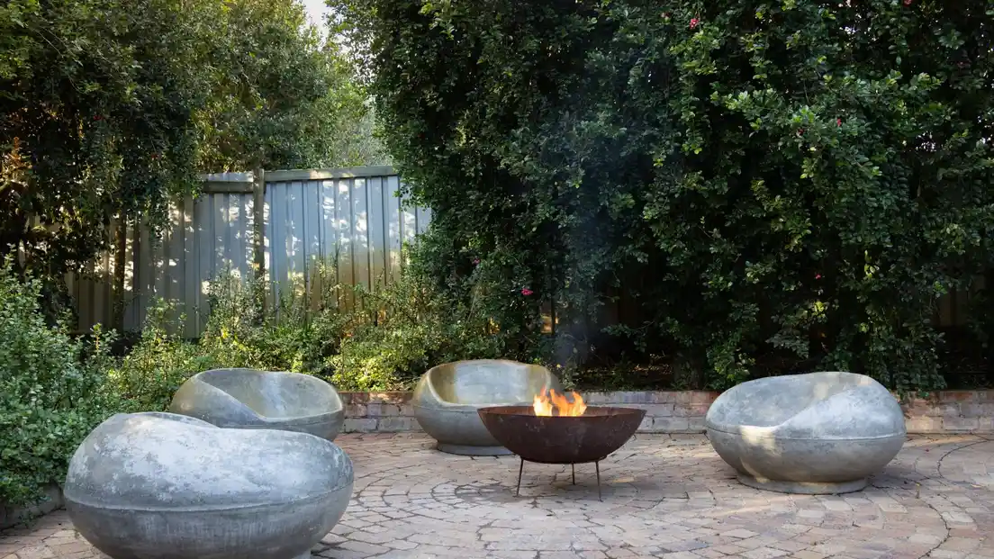 A lit corten steel fire bowl on a reclaimed-brick paver patio surrounded by greenery