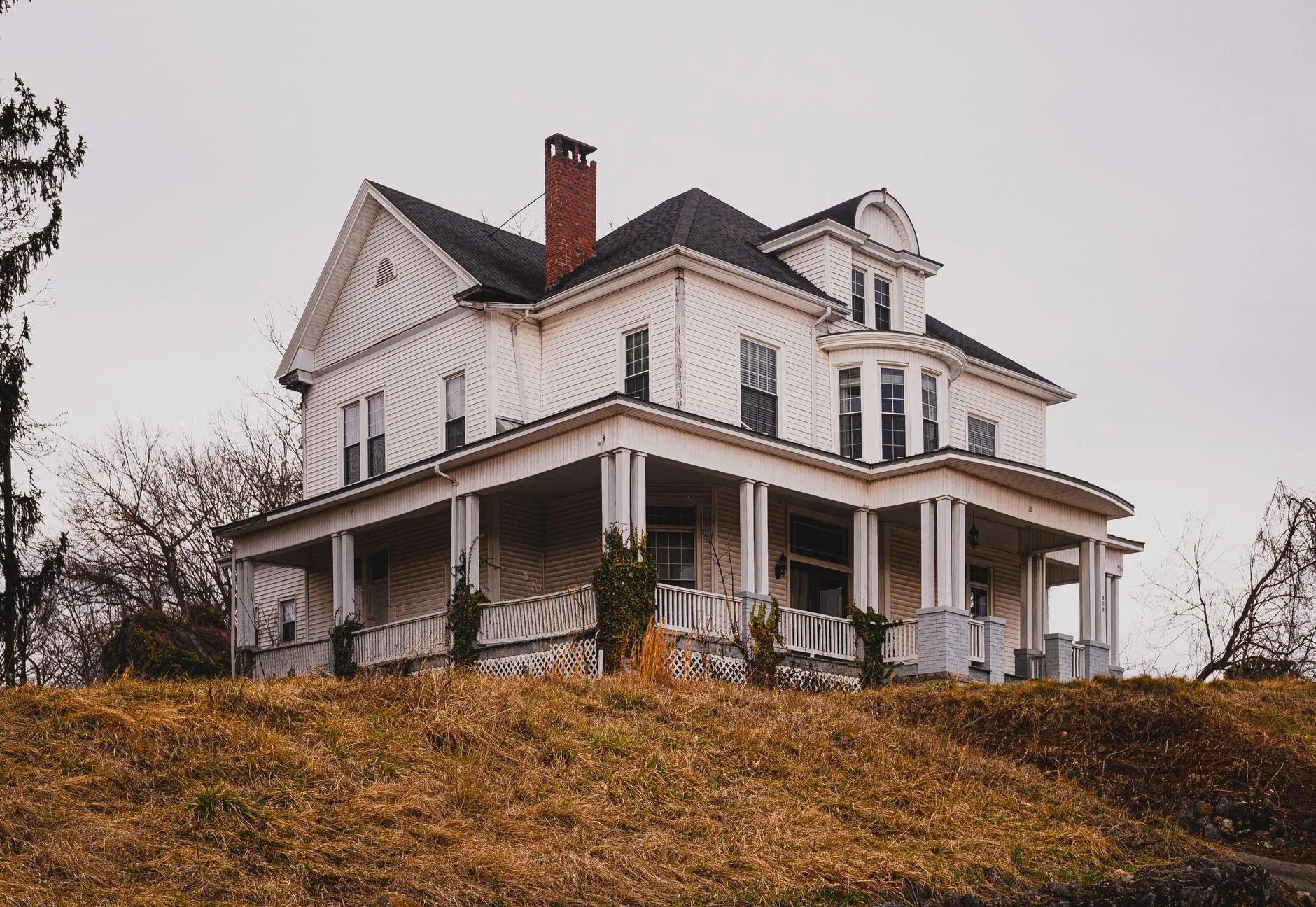 A weathered traditional white farmhouse with a wraparound porch, contrasting a modern farmhouse exterior