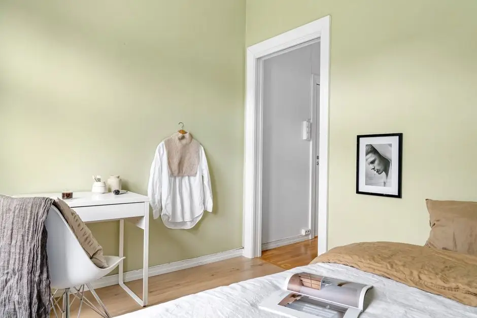 A bedroom with soft sage green walls and a small white desk, an example of a calming bedroom wall color for a room with good natural light