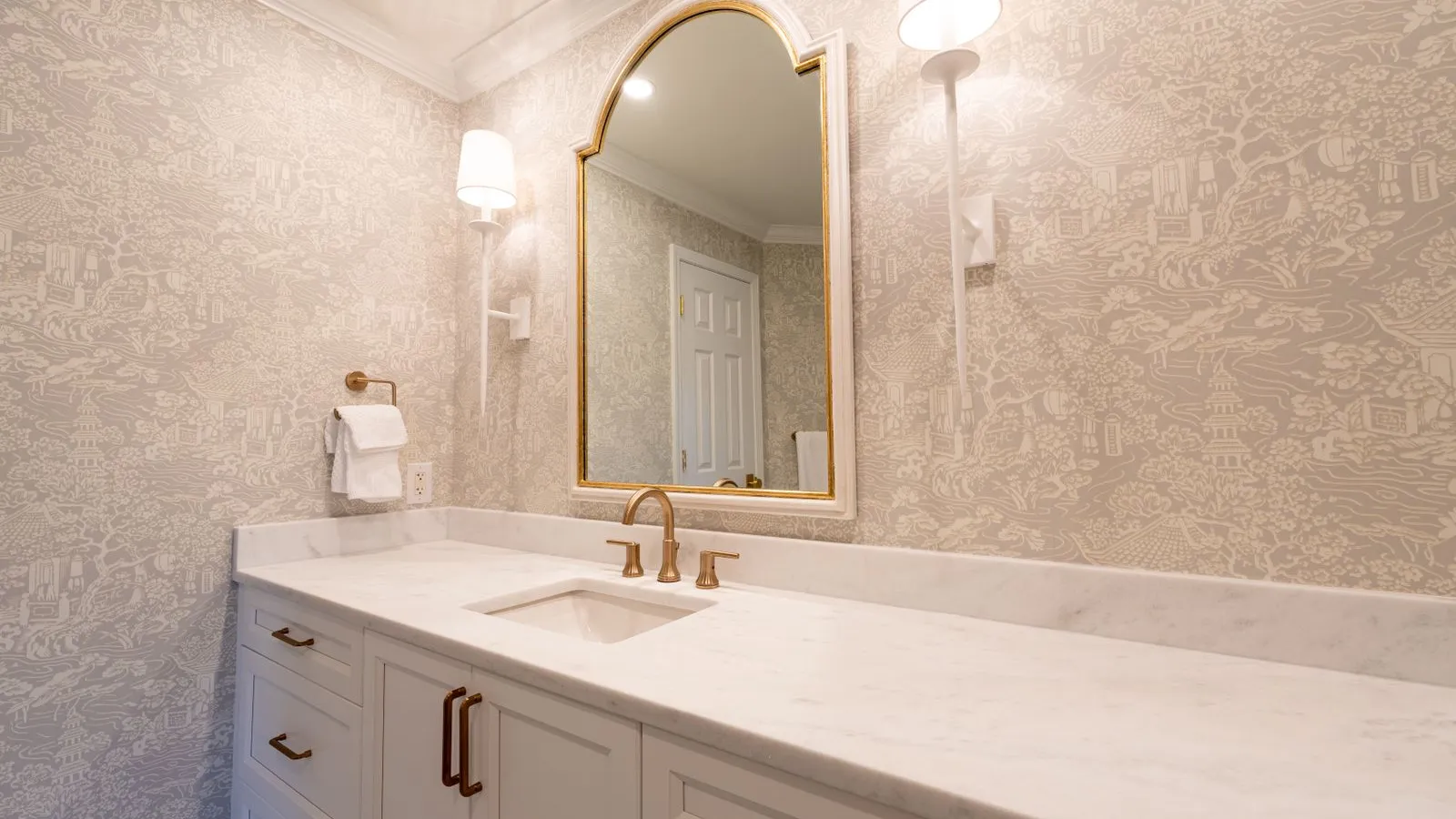 An arched gold-framed mirror flanked by two lit wall sconces above a marble vanity with a wallpapered wall
