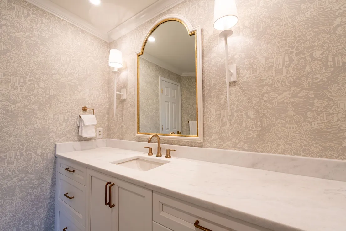An arched gold-framed mirror flanked by two lit wall sconces above a marble vanity with a wallpapered wall