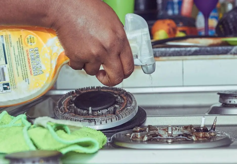A hand cleaning a gas stove burner with spray cleaner and a cloth