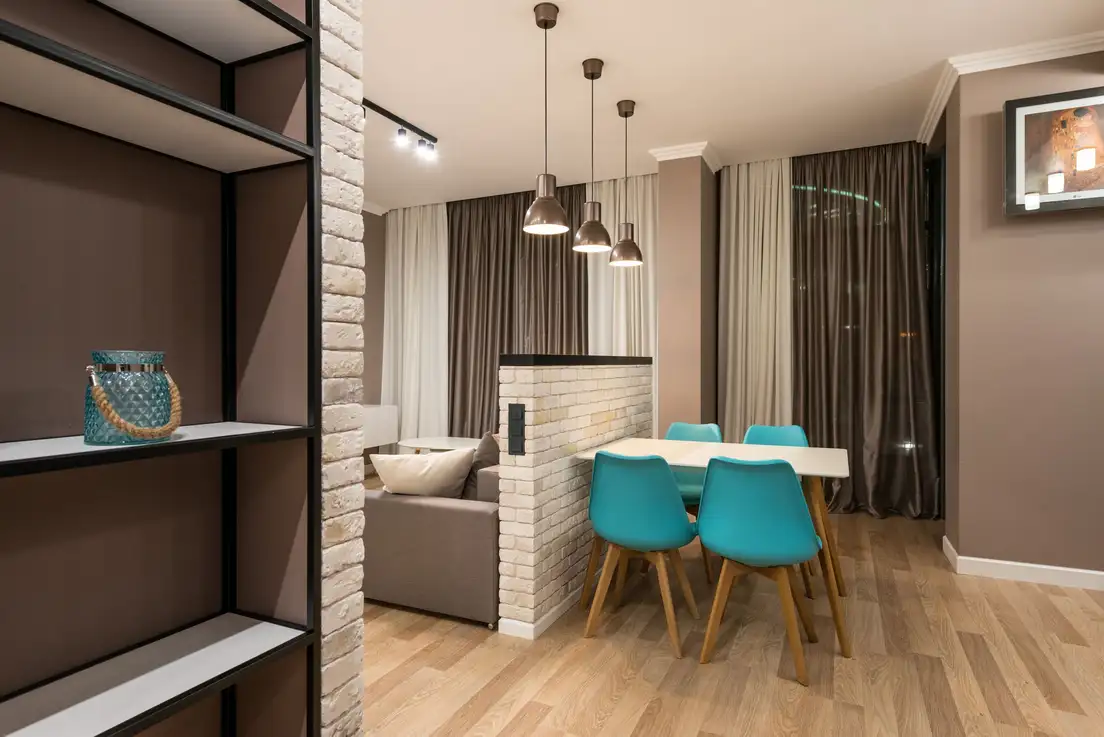 Open metal shelving and a half brick wall separating a dining table with blue chairs from a sofa in a small living room