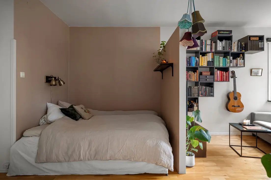 A small bedroom with a bed pushed into a corner against a blush wall, next to a bookshelf divider and living area