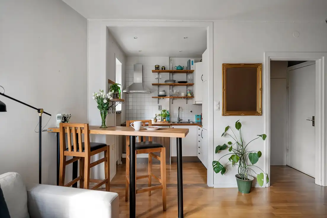 A small living room with warm white walls, a natural wood counter with wooden stools, a gold-framed mirror, and a potted plant