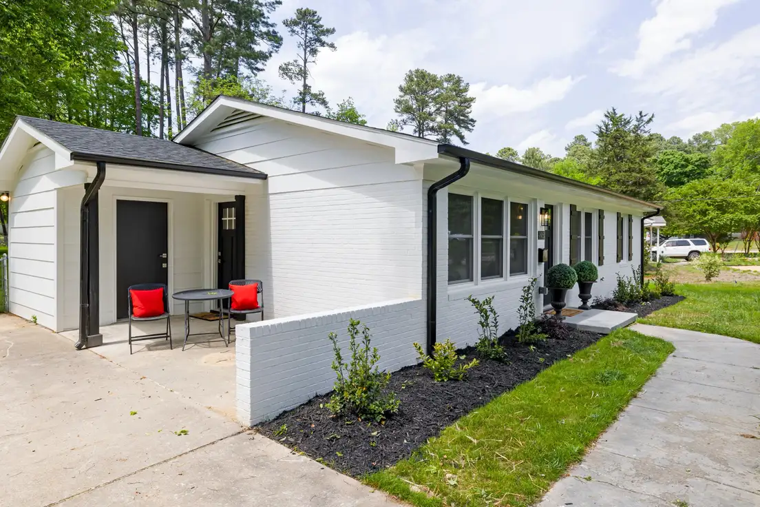 A small concrete front porch on a white brick ranch house with a bistro table and two black chairs with red cushions