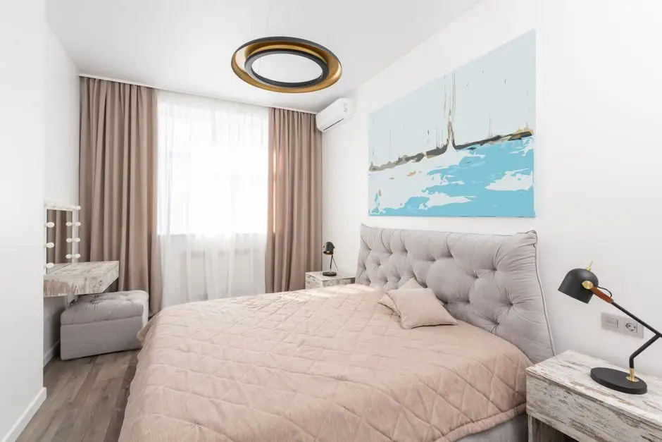 A small bedroom in light neutral wall tones with a tufted headboard and blue artwork, an example of the best wall color for a small bedroom