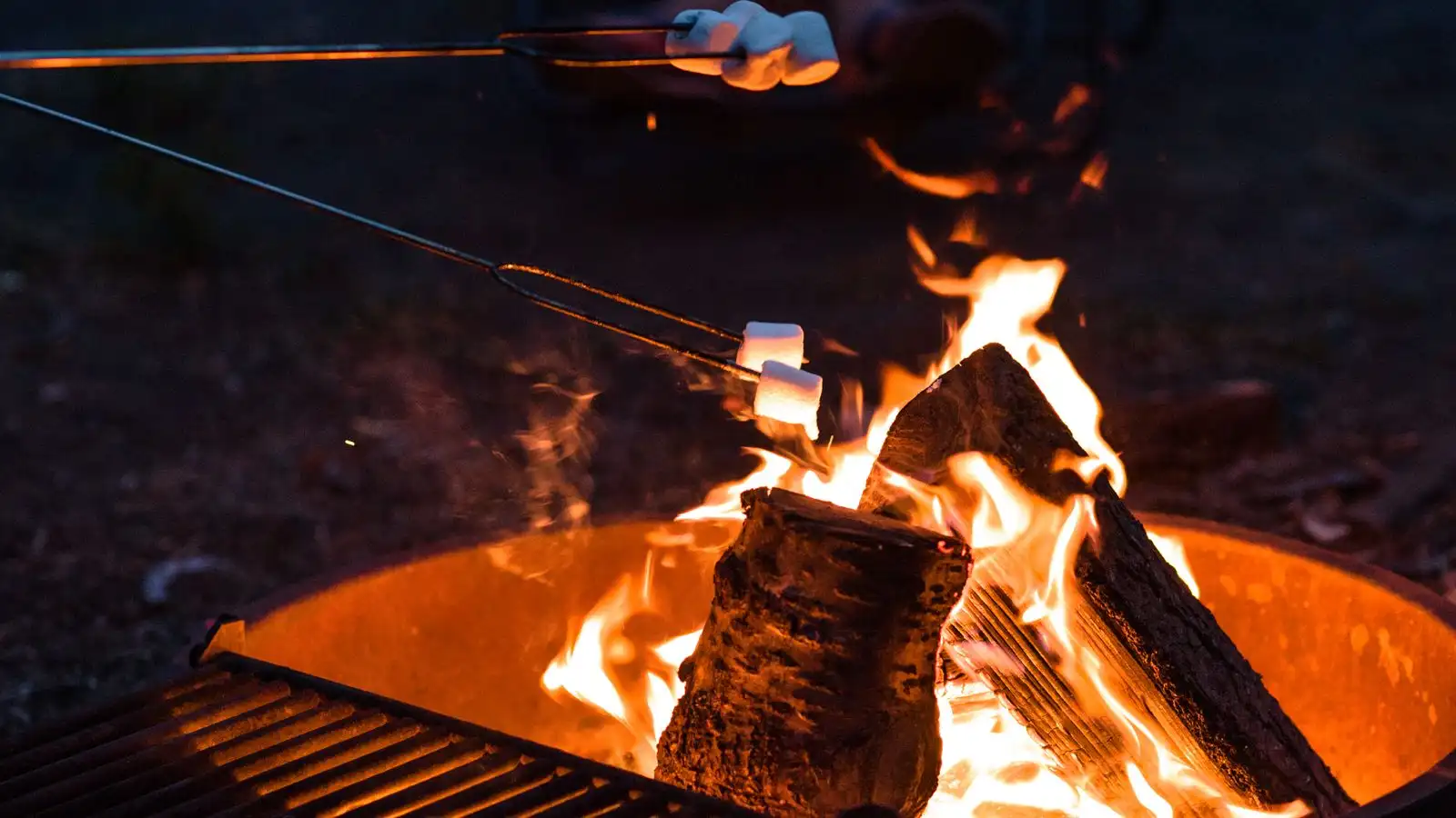 A marshmallow on a roasting stick held over a rusty steel backyard fire pit at dusk