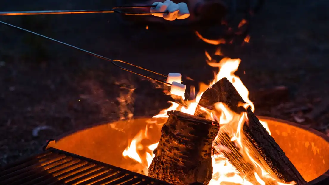 A marshmallow on a roasting stick held over a rusty steel backyard fire pit at dusk