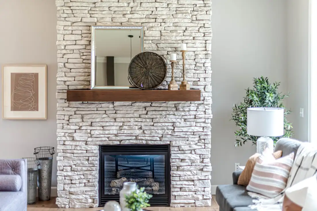 A white stacked-stone fireplace with a dark wood mantel shelf styled with a mirror, woven tray, and two wooden candlesticks