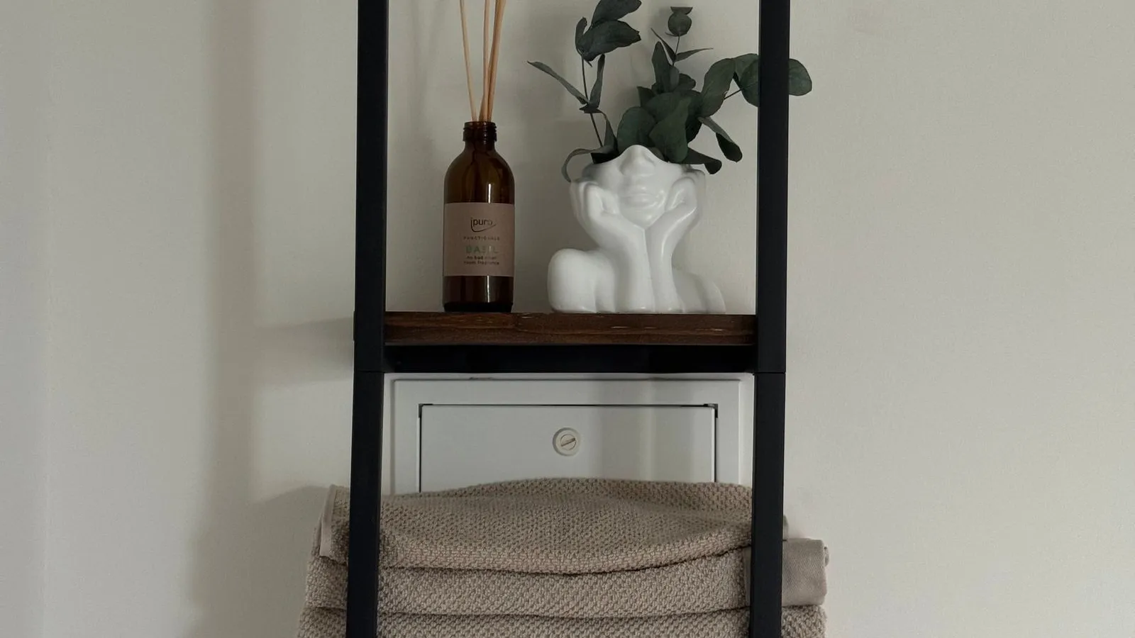 A black wall-mounted ladder shelf holding folded towels, a diffuser, and a small vase