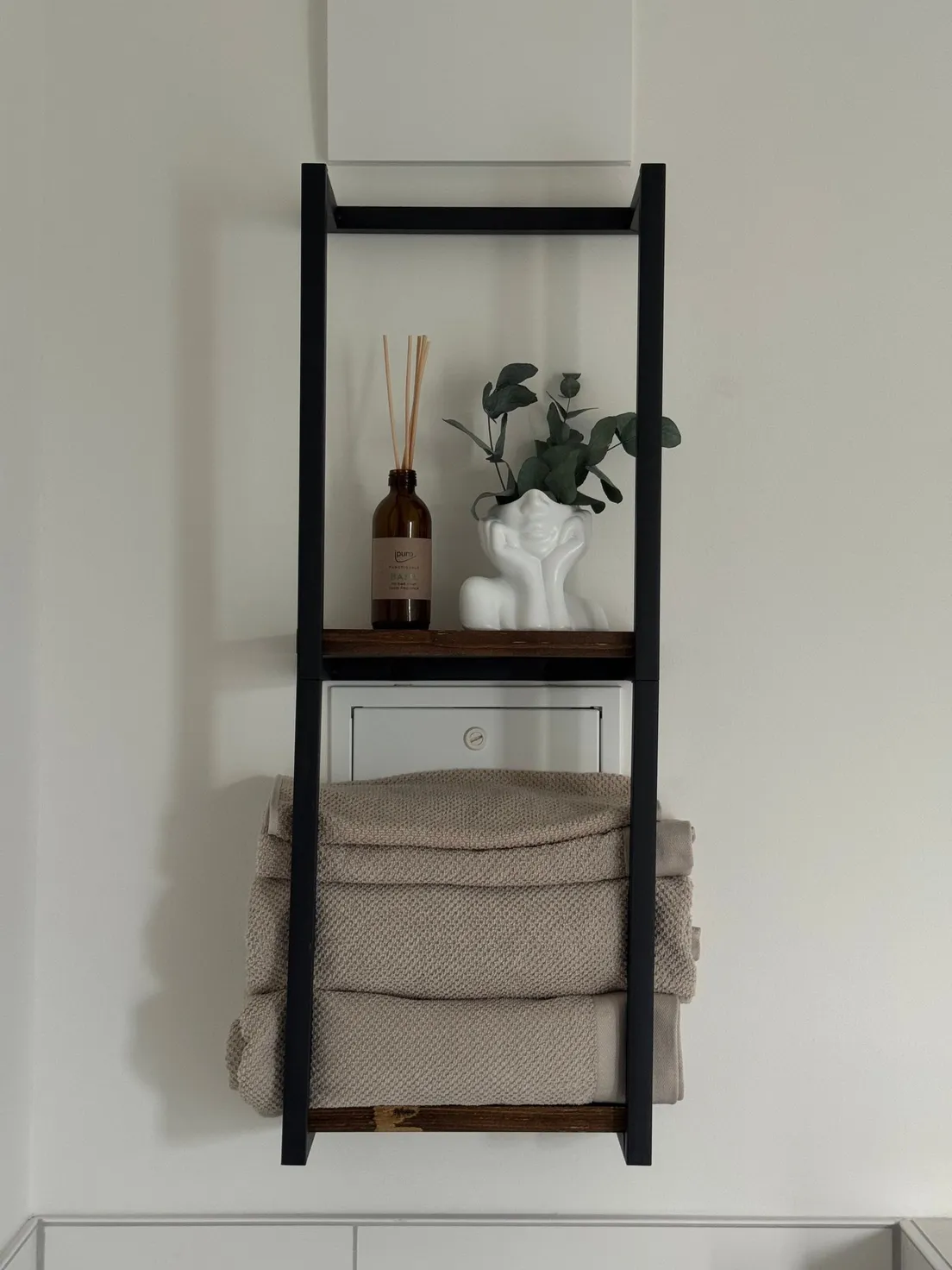 A black wall-mounted ladder shelf holding folded towels, a diffuser, and a small vase