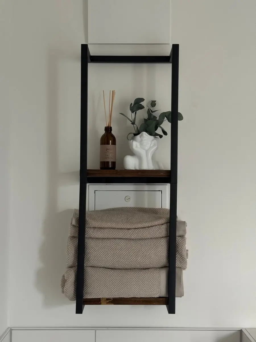 A black ladder shelf mounted above a bathroom wall holding folded towels and decor
