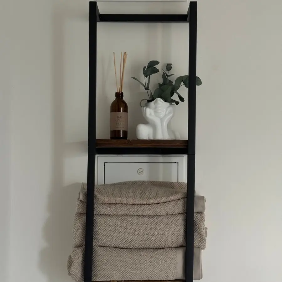 A black ladder shelf mounted above a bathroom wall holding folded towels and decor