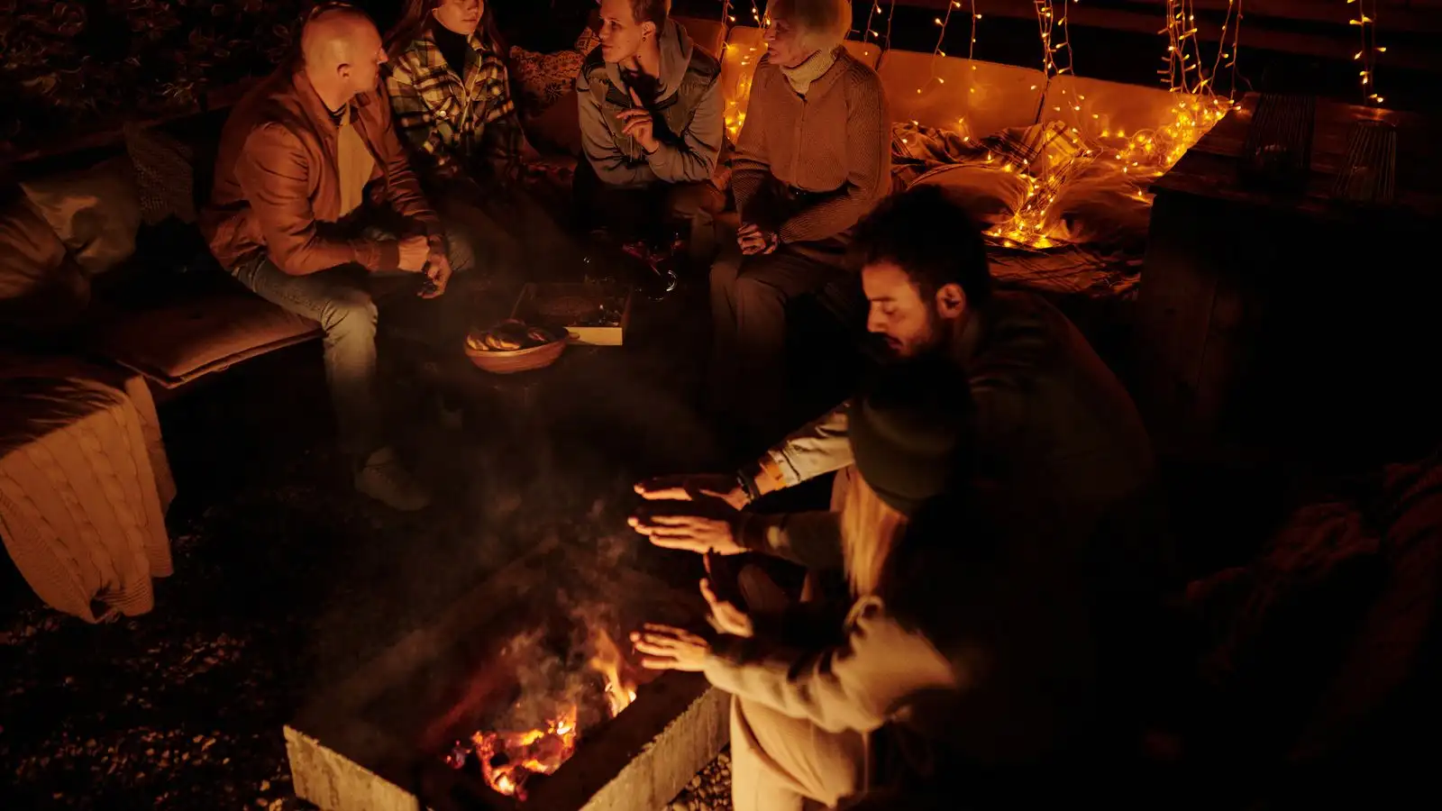 Friends gathered around a backyard fire pit at night under warm string lights strung along the fence