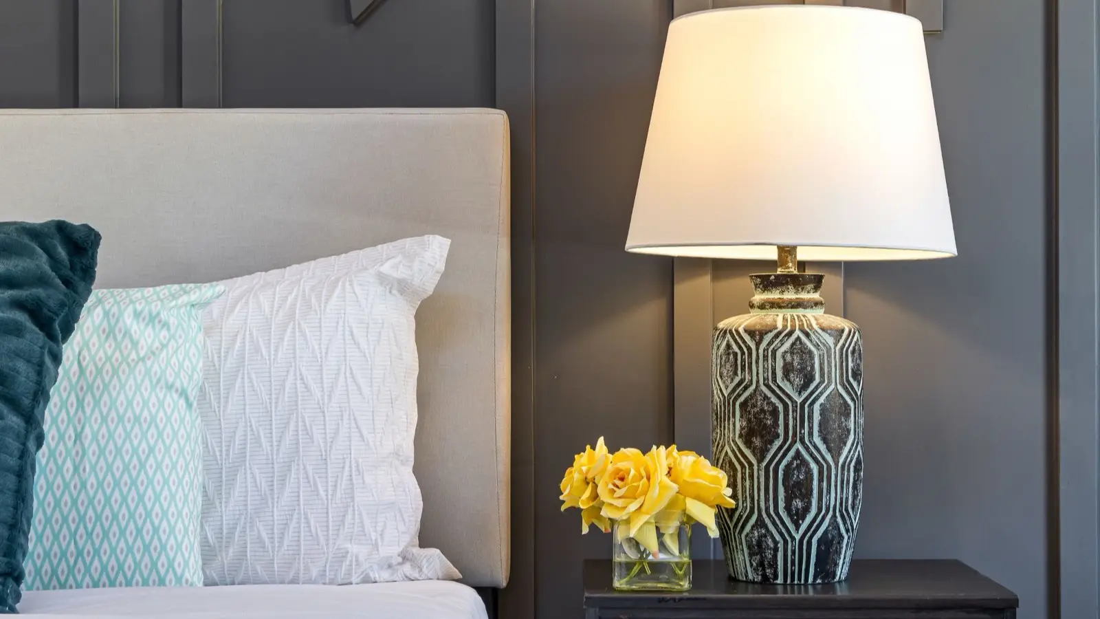 A styled bedside table with a patterned ceramic lamp and fresh yellow roses in a glass vase