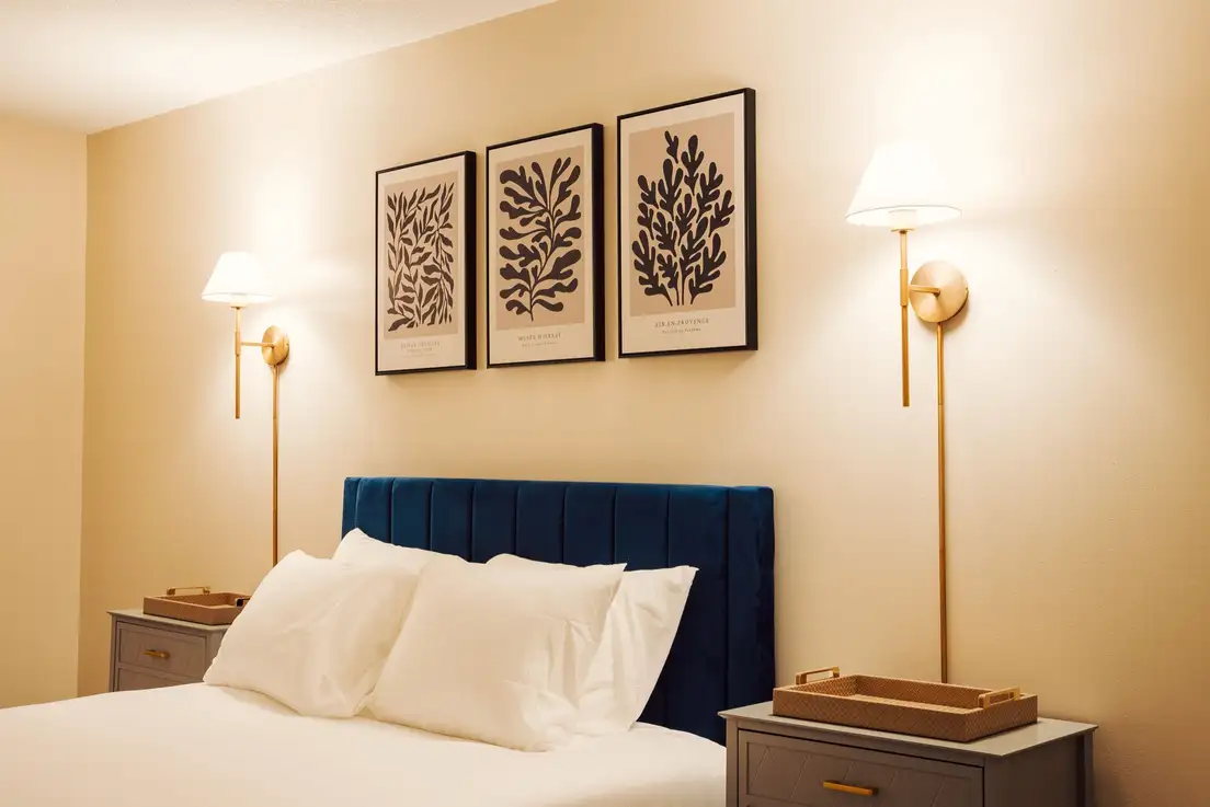 Two brass plug-in wall sconces with white shades lighting each side of a bed with a navy velvet headboard