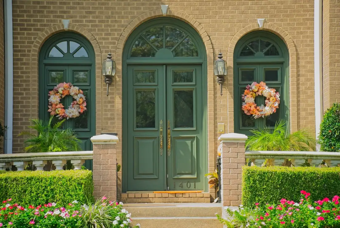 A symmetrical brick entry with green arched doors, matching lanterns, matching wreaths, and trimmed hedges on each side