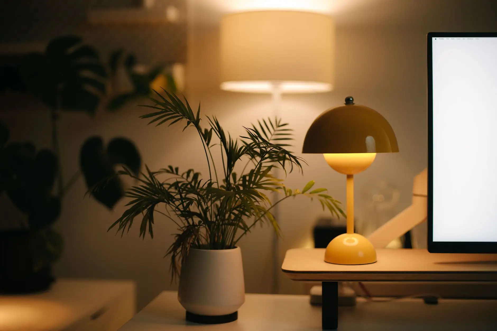 A warm mushroom table lamp glowing on a desk beside a potted palm and a monitor in a living room workspace