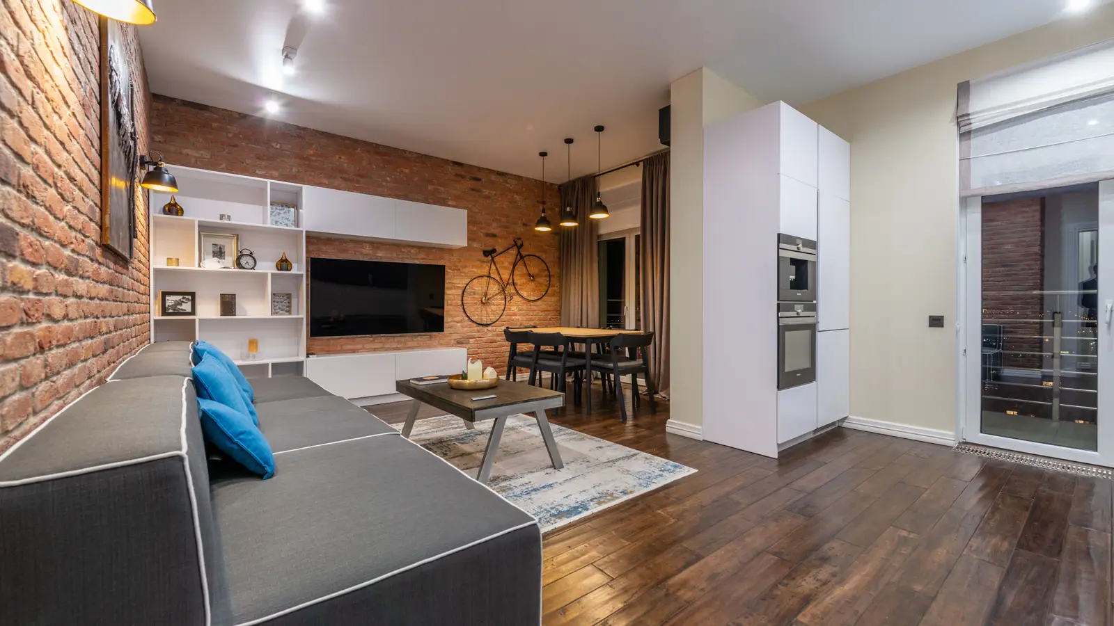 A wall-mounted TV against an exposed brick accent wall next to white built-in shelving