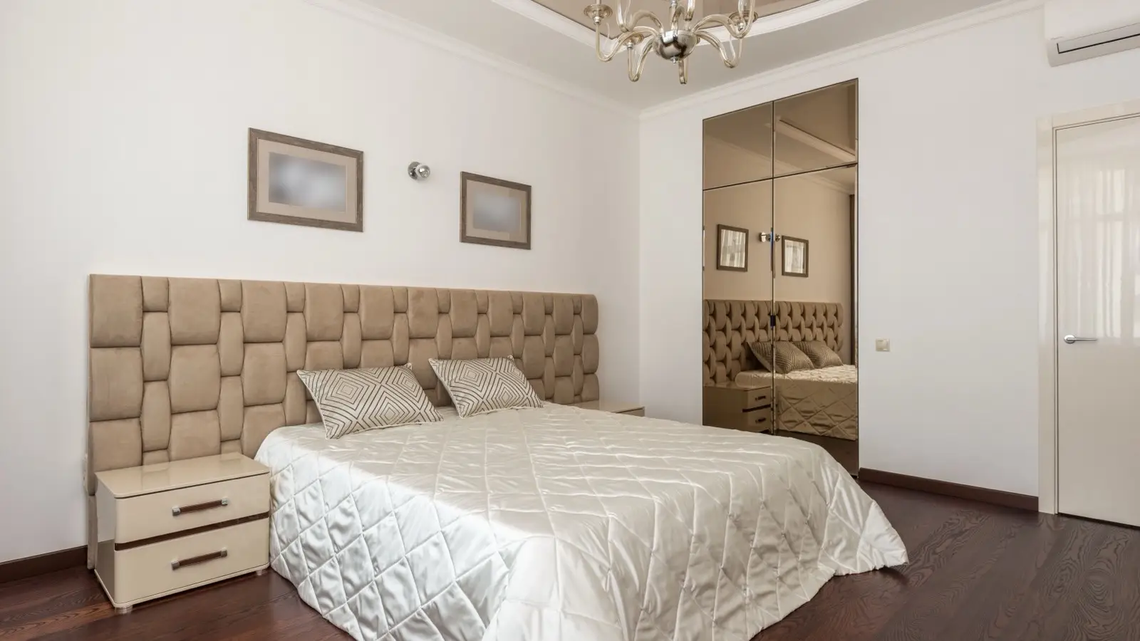 A beige diamond-tufted headboard with a cream quilted bedspread and matching nightstand
