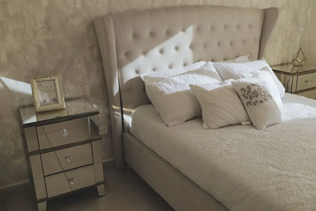 A queen bed with a tufted wingback headboard, layered white pillows, and a mirrored nightstand in a shared bedroom