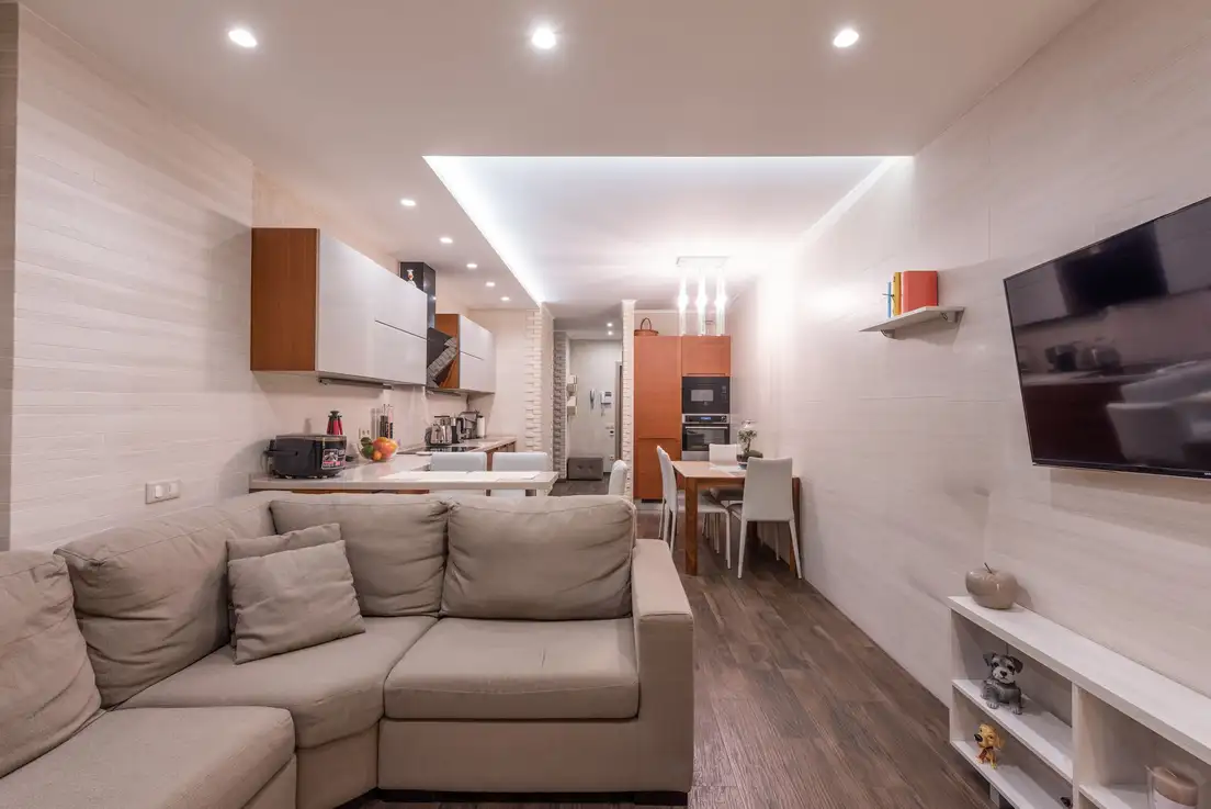 A long rectangular living room with a sectional sofa facing a wall-mounted TV and a dining table at the far end near the kitchen