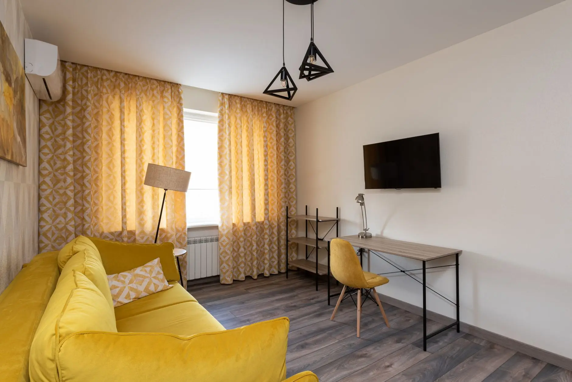 A slim black metal desk with a yellow chair sharing a wall with a mounted TV in a small living room with a yellow sofa