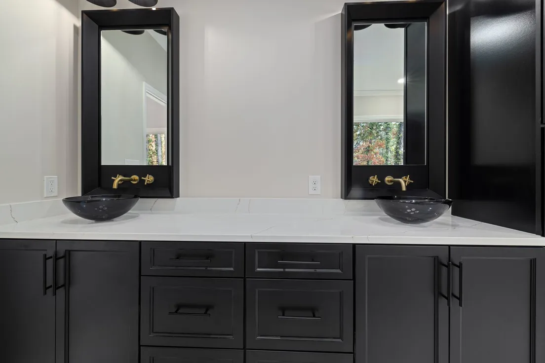 A dark bathroom vanity with twin glass vessel sinks and brass wall-mounted faucets against a black marble backsplash