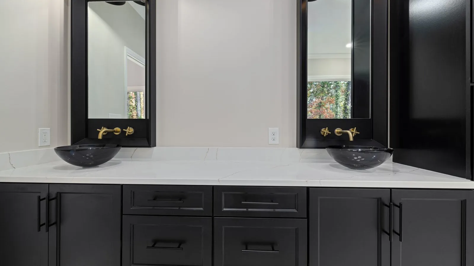 A dark bathroom vanity with twin glass vessel sinks and brass wall-mounted faucets against a black marble backsplash
