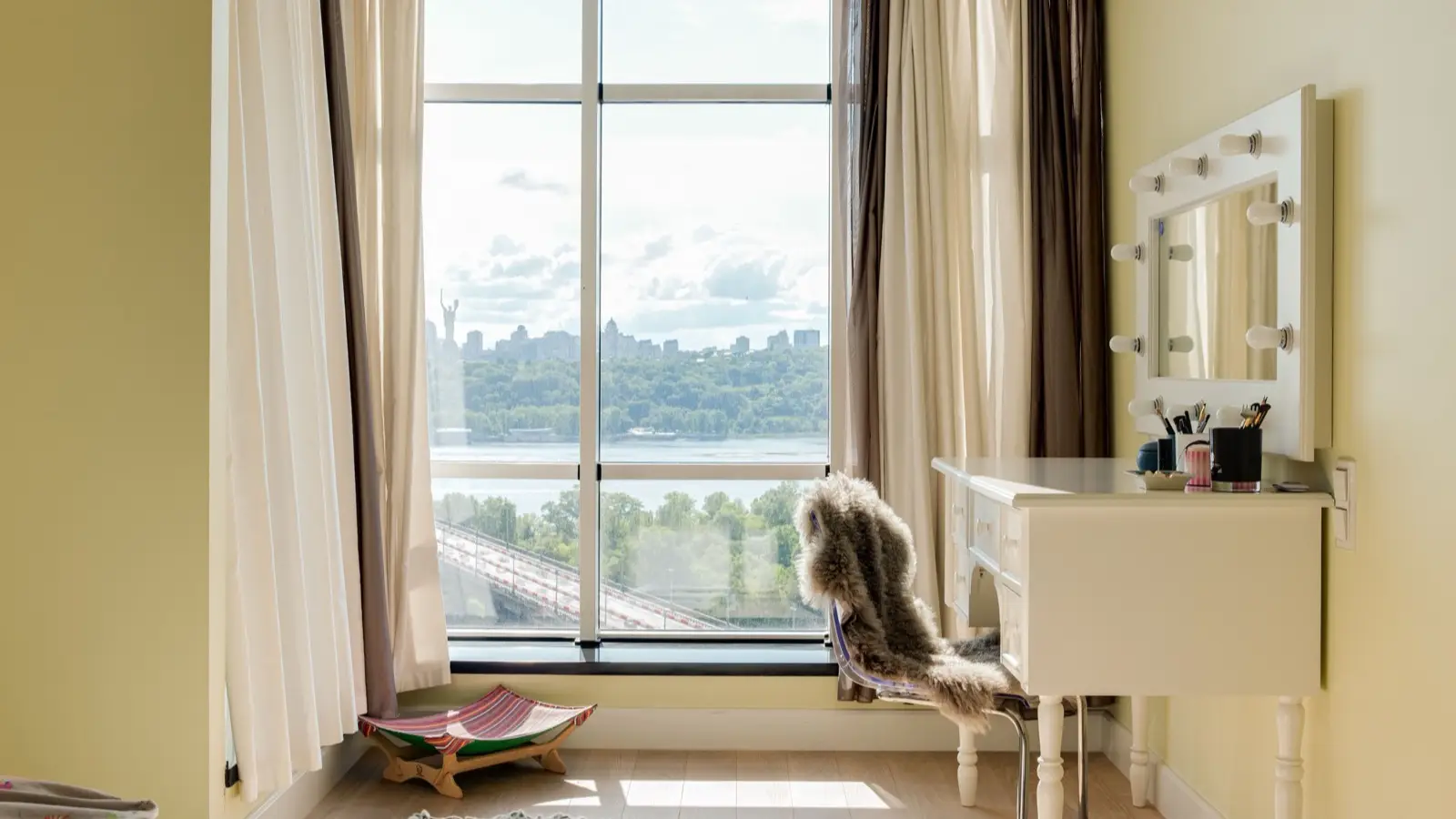 A white vanity table with a Hollywood-style lit mirror, a fur throw, and a city view window