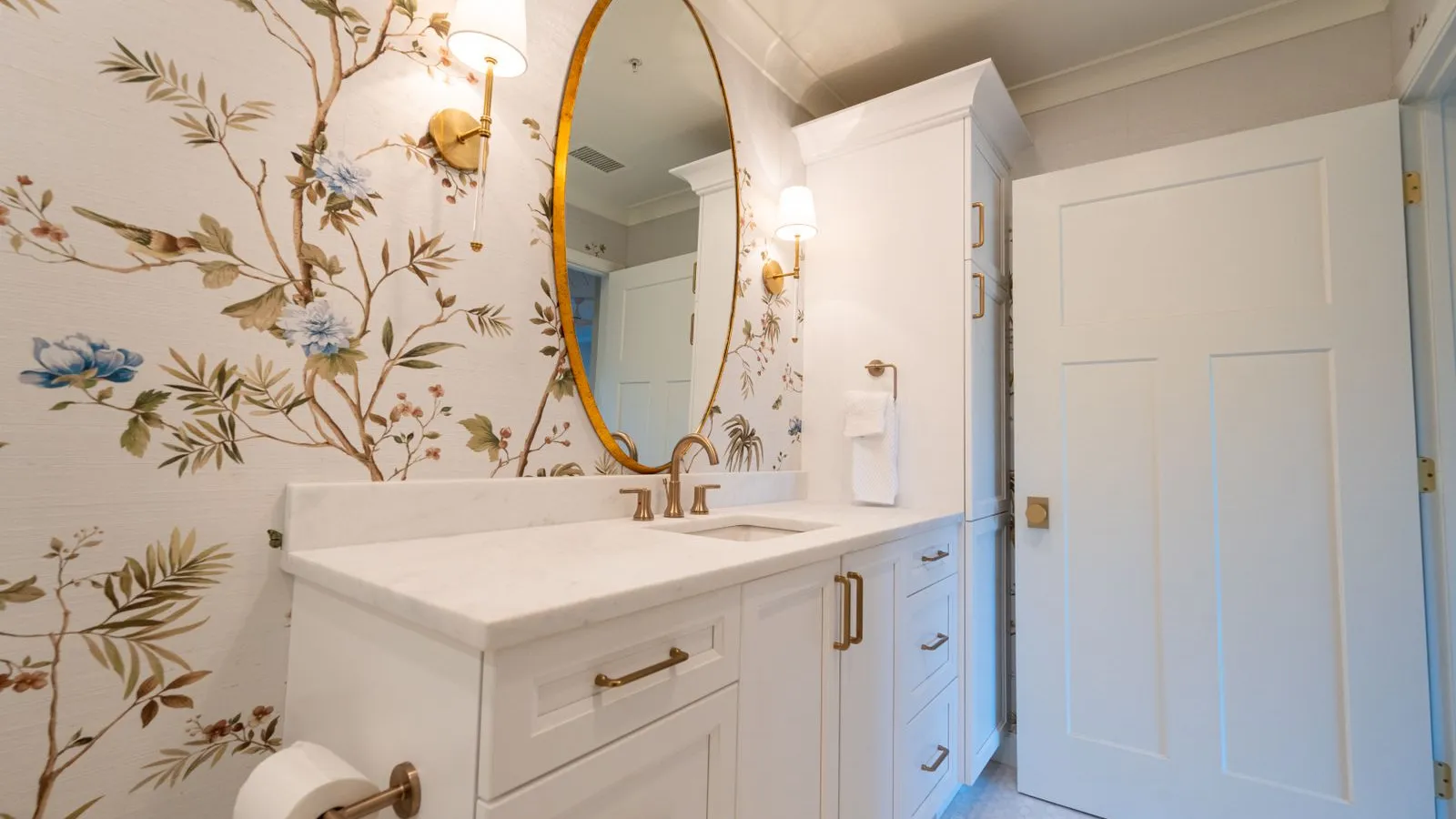 A powder room with bold floral wallpaper on every wall, an oval brass-framed mirror, and gold sconces