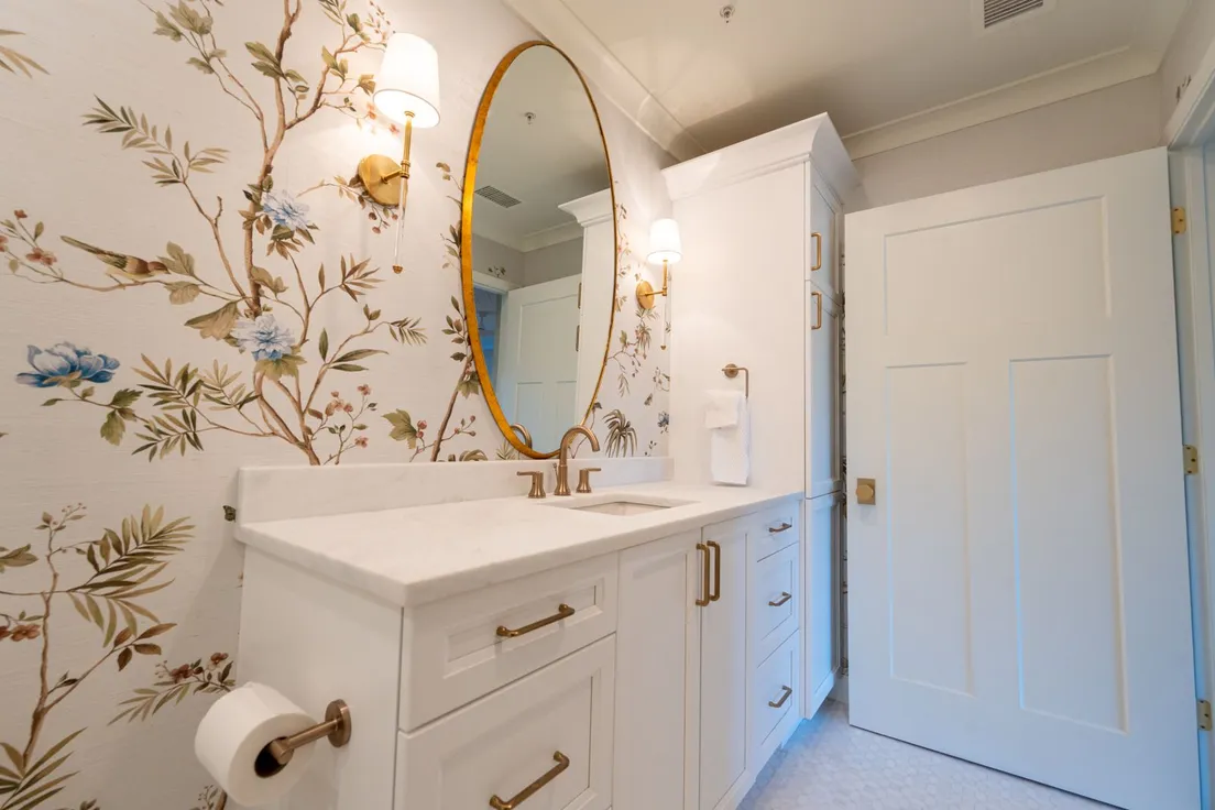 A powder room with bold floral wallpaper on every wall, an oval brass-framed mirror, and gold sconces
