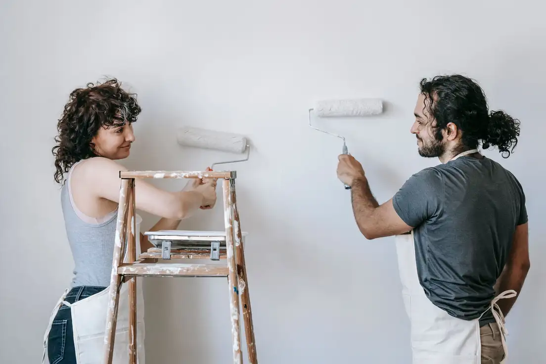 Two people rolling warm white paint onto a living room wall from a stepladder, the first step of a budget farmhouse makeover