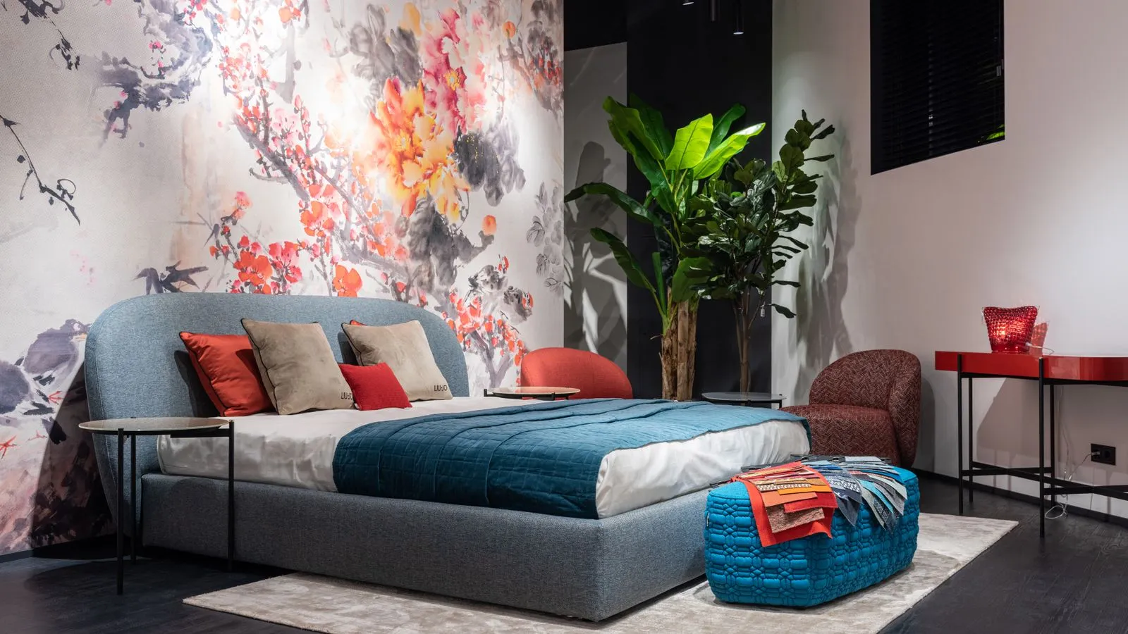 A colorful bedroom with a bold floral wall mural, coral and blue cushions, a teal bedspread, and large plants