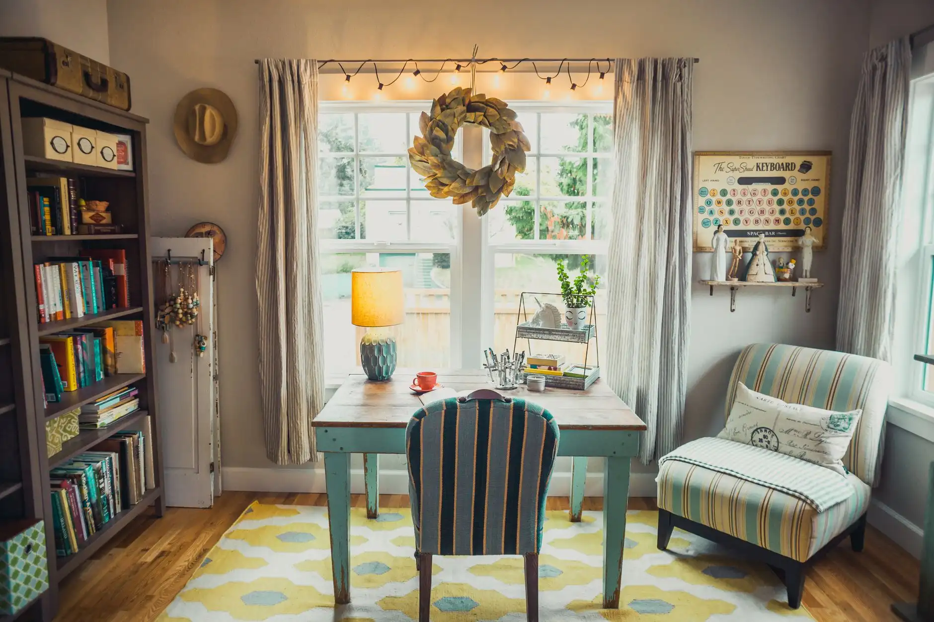 A painted wood desk facing a window with an upholstered chair, bookcase, and armchair in a small living room workspace