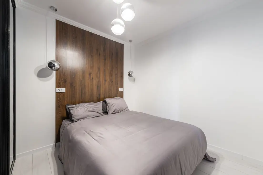 A dark walnut wood panel feature wall behind a low bed with grey bedding and chrome pendant lights