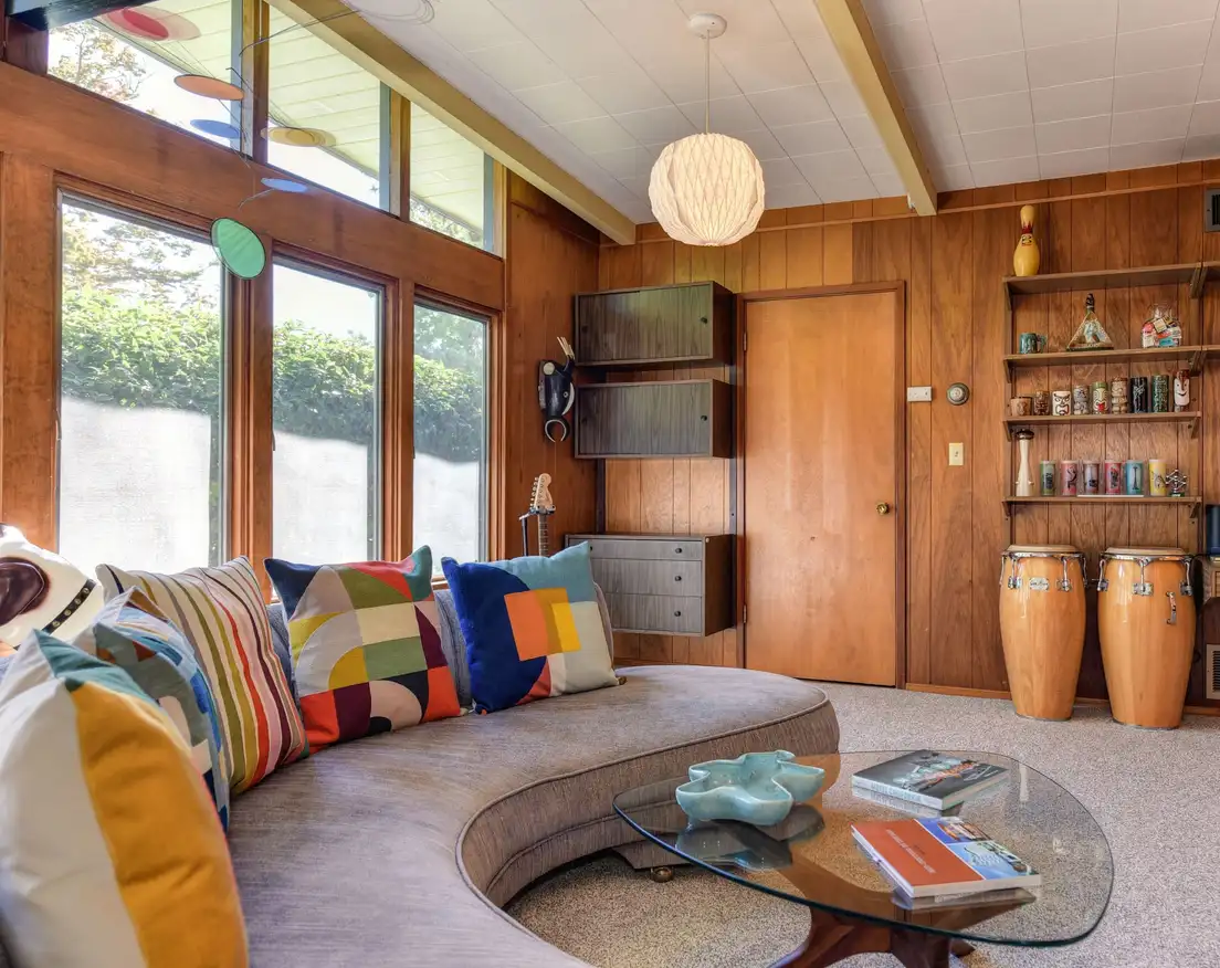 A mid-century ranch den with warm wood paneling, clerestory windows above tall windows, a curved sofa, and a paper globe pendant