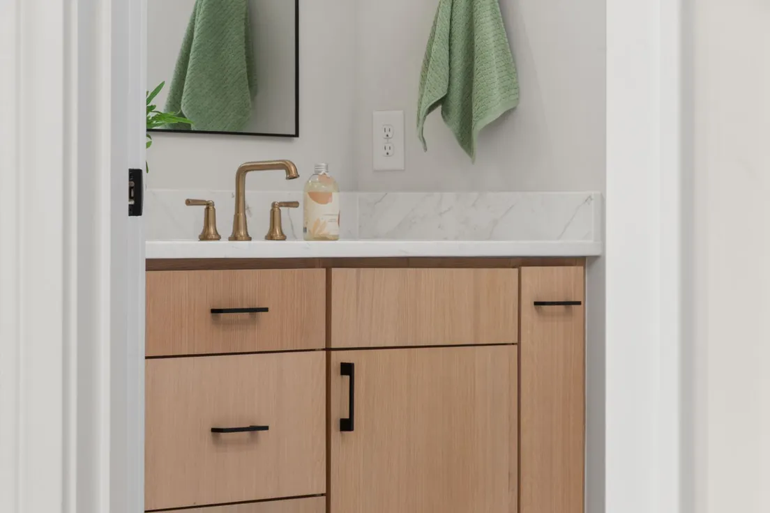 A light wood bathroom vanity with brass faucet and hardware beneath a mirror, with green towels hanging on the wall