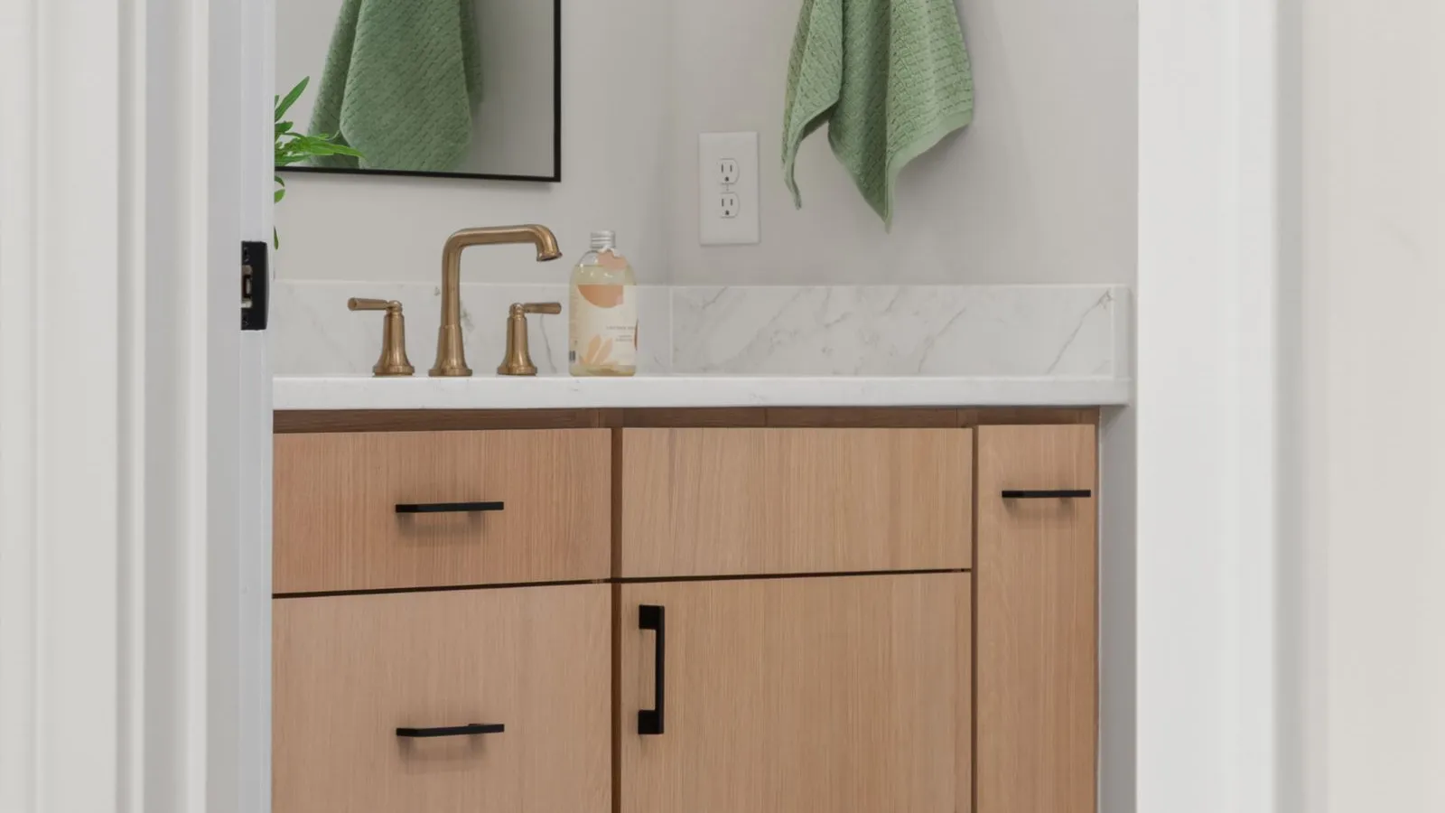 A light wood bathroom vanity with brass faucet and hardware beneath a mirror, with green towels hanging on the wall